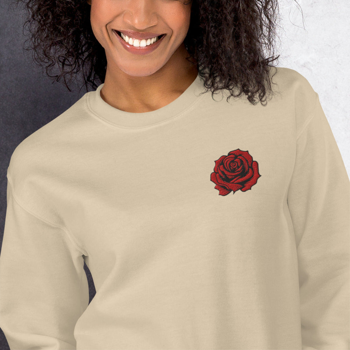 Front view of a sand colored women's crewneck sweatshirt featuring a red embroidered rose on the chest. The warm neutral shade and delicate floral stitching create a versatile look that pairs well with denim, casual trousers, and leggings. This embroidered rose sweatshirt is a thoughtful gift for women who enjoy understated floral clothing and classic botanical designs.
