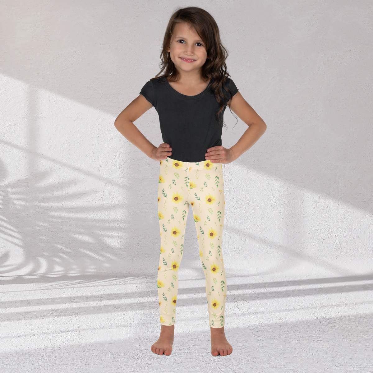 Girls leggings featuring a cheerful wildflower inspired print with yellow sunflowers, green leaves, delicate stems and small flowers on a soft cream background. The full length leggings have a high rise waistband and a smooth stretchy fabric designed for comfortable everyday movement. The botanical pattern has a pretty cottagecore garden feeling that works for school, playtime, spring outfits, vacations, family photos and casual days outside.