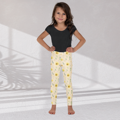 Girls leggings featuring a cheerful wildflower inspired print with yellow sunflowers, green leaves, delicate stems and small flowers on a soft cream background. The full length leggings have a high rise waistband and a smooth stretchy fabric designed for comfortable everyday movement. The botanical pattern has a pretty cottagecore garden feeling that works for school, playtime, spring outfits, vacations, family photos and casual days outside.