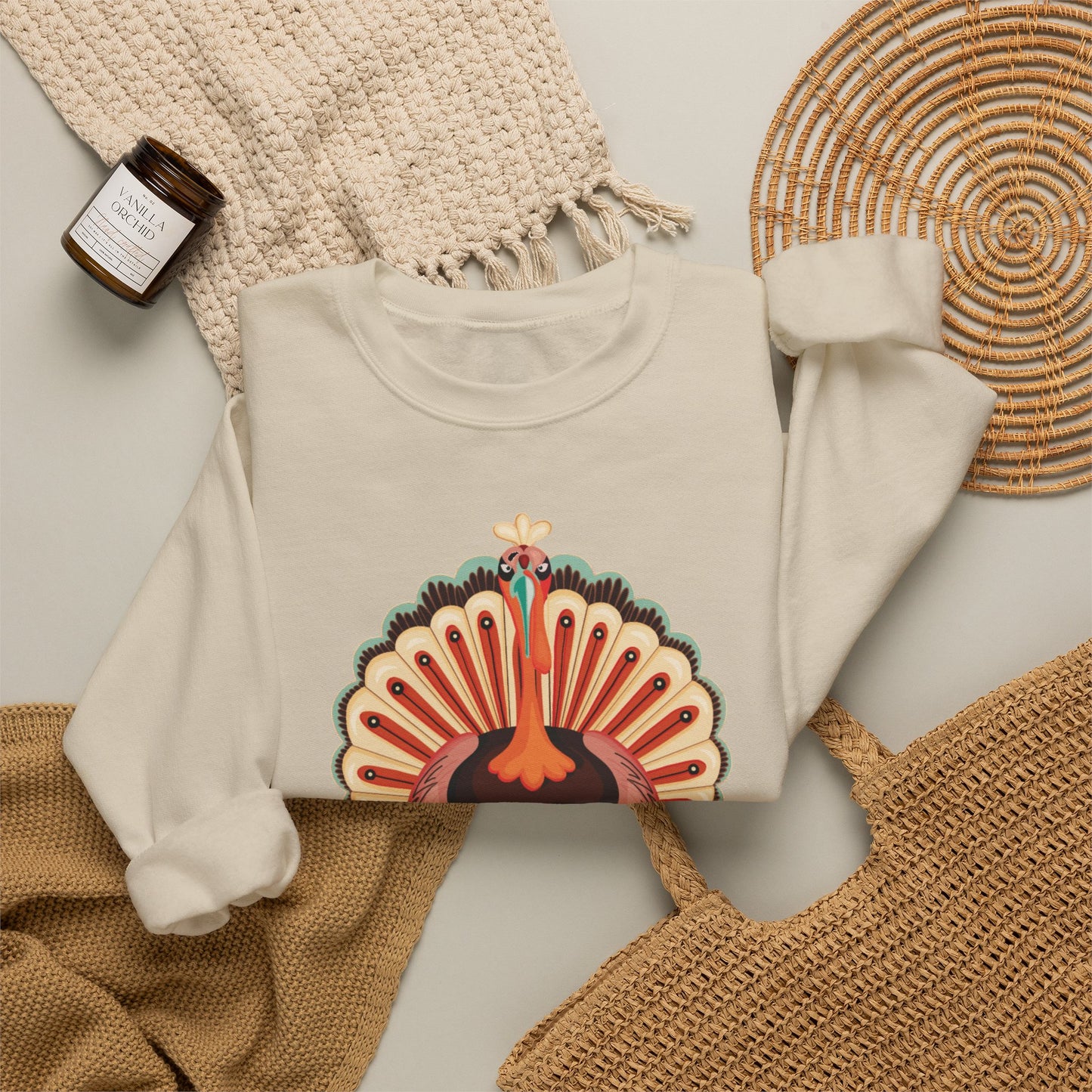 An off-white crewneck sweatshirt with a colorful graphic of a thanksgiving turkey with its tail feathers fanned, styled in a flat lay with a woven blanket, a small candle, and a wicker tray.