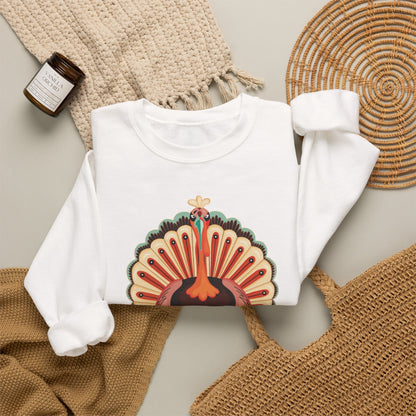 A white long-sleeve crewneck sweatshirt laid flat on a neutral background, featuring a colorful graphic of a turkey with an open tail. The sweatshirt is styled with a beige knit blanket, a woven straw bag, a round woven placemat, and a small amber candle jar.