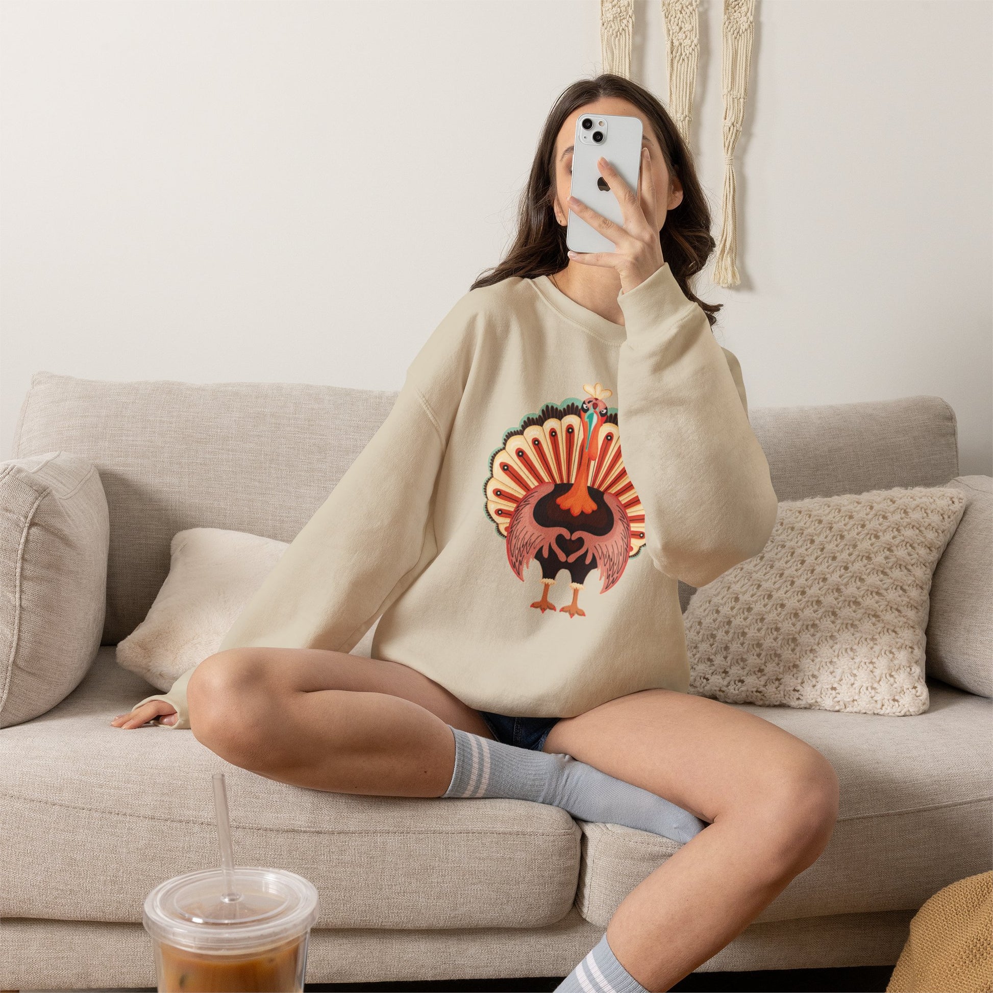 A woman takes a mirror selfie while sitting on a beige couch, wearing a cream-colored sweatshirt with a colorful turkey graphic on the front. She is wearing gray striped socks and has an iced coffee in the foreground. Thanksgiving turkey sweatshirt with adorable colorful turkey making a heart shape with its wings, a cute family matching outfit for Thanksgiving dinner and fall gatherings