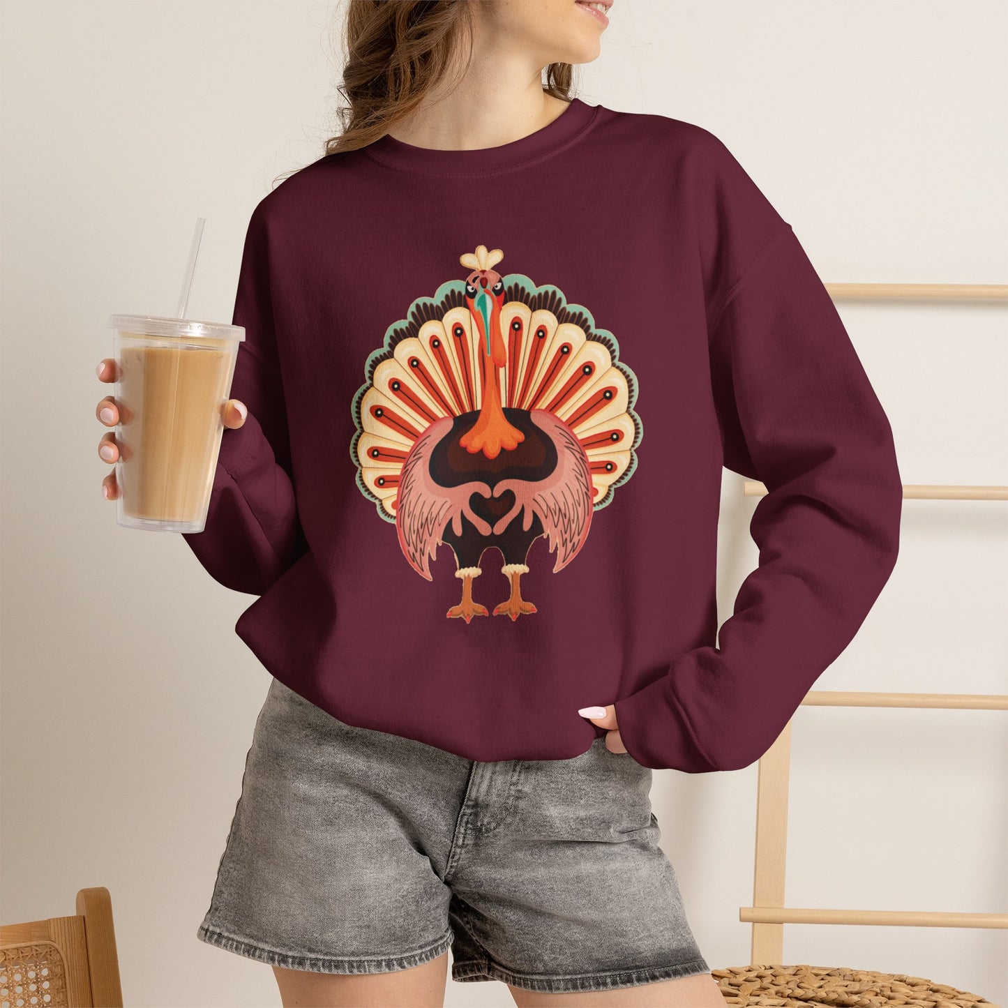 A woman wearing a maroon crewneck sweatshirt with a colorful graphic of a stylized turkey, holding an iced coffee in a bright indoor setting. Cute Thanksgiving turkey sweatshirt featuring a colorful cartoon turkey with its wings forming a heart, perfect for family matching Thanksgiving outfits and festive fall celebrations
