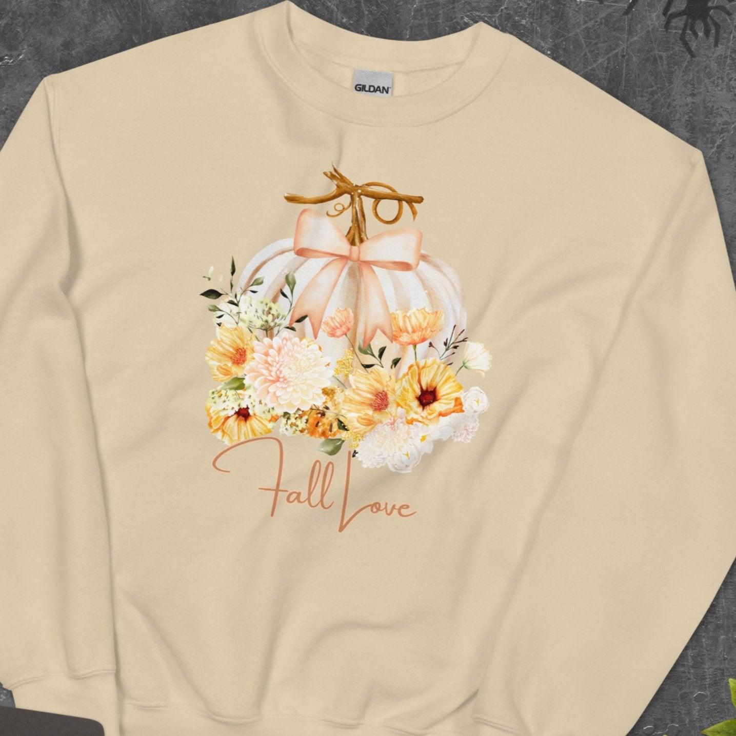 Fall Love Ladies Sweatshirt, this is a Sand colored Gildan Sweatshirt, with a graphic of a white pumpkin with a peach ribbon. The pumpkin is surrounded by a garland of fall leaves and flowers, with the text- Fall Love, by jaecrece