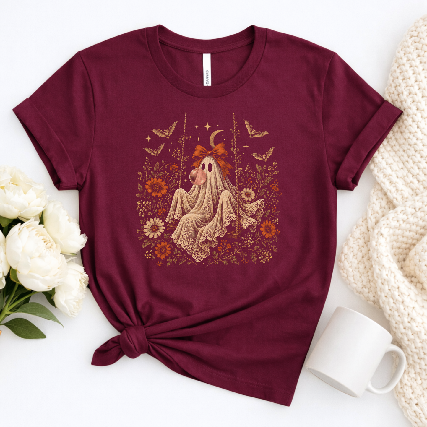 Maroon girls Halloween T shirt shown in a flatlay featuring a whimsical vintage ghost blowing bubble gum, surrounded by orange flowers, botanical foliage, bats, stars, lace and a bow. A cute spooky season graphic tee for girls who love vintage Halloween artwork, fall fashion and playful ghost designs.