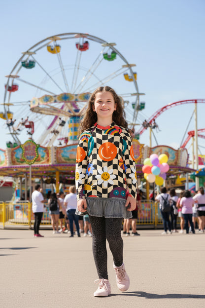 Tween girl walking through an amusement park wearing a retro wavy checkerboard girls rash guard with colorful smiley faces, rainbows, daisies, stars, and swirls. Long sleeve UPF 50+ swim shirt available in sizes 2T–20.