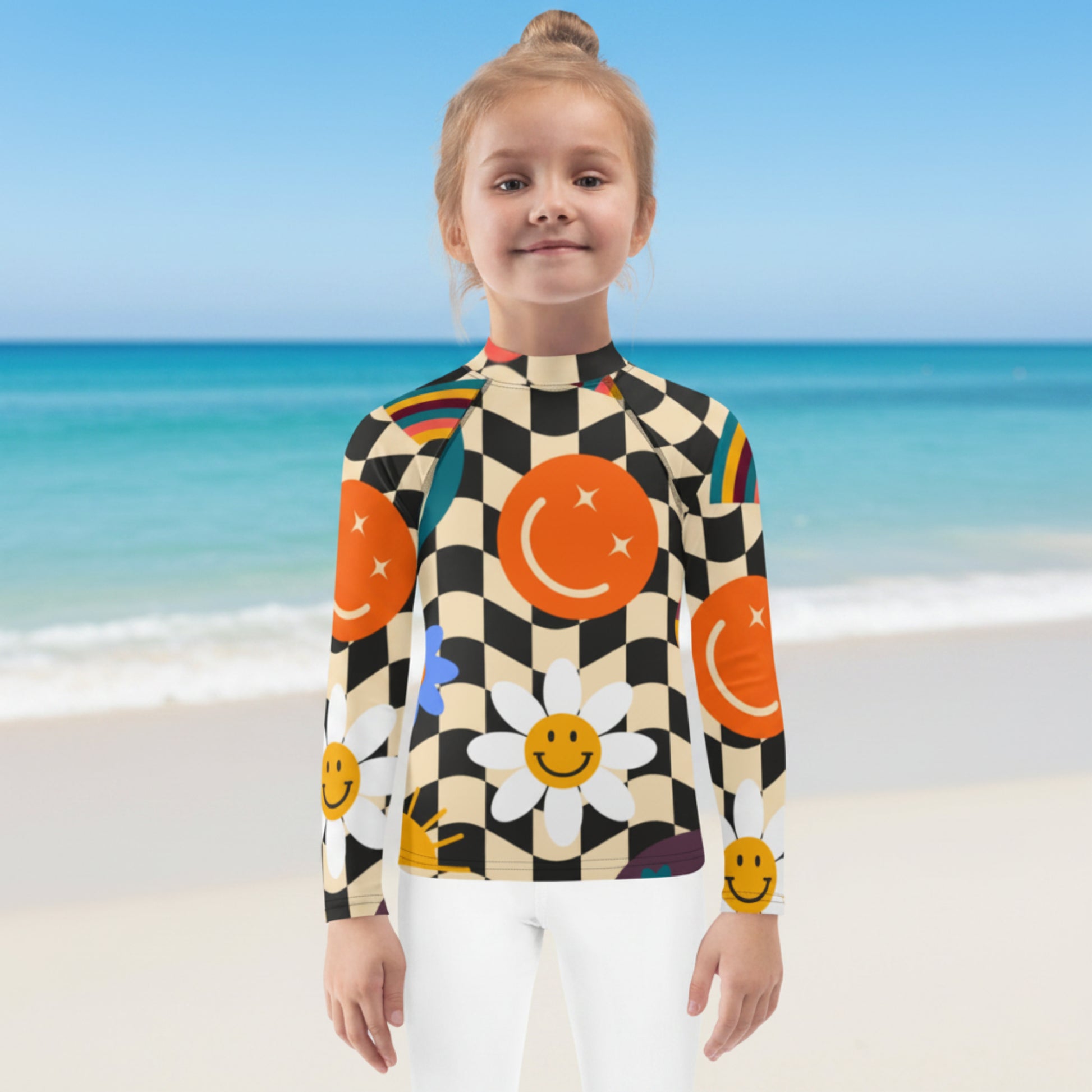 Young girl enjoying a beach vacation while wearing a retro wavy checkerboard girls rash guard with colorful smiley faces, rainbows, daisies, stars, and swirls. Long sleeve UPF 50+ swim shirt available in sizes 2T–20 with coordinating family swimwear.