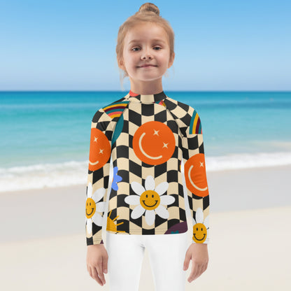 Young girl enjoying a beach vacation while wearing a retro wavy checkerboard girls rash guard with colorful smiley faces, rainbows, daisies, stars, and swirls. Long sleeve UPF 50+ swim shirt available in sizes 2T–20 with coordinating family swimwear.