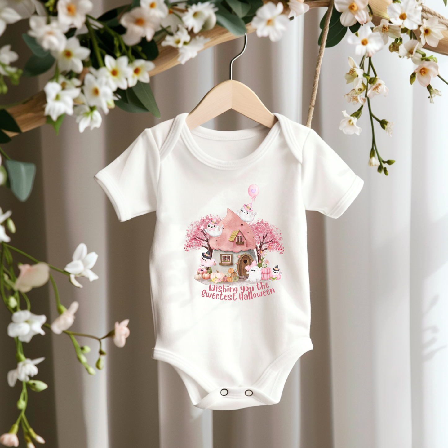 A white short-sleeve baby bodysuit hanging on a wooden hanger, featuring a cute Halloween graphic of a haunted house, pumpkins, and the text 'Wishing you the Sweetest Halloween'. The bodysuit is styled against a soft curtain backdrop with hanging white flowers.