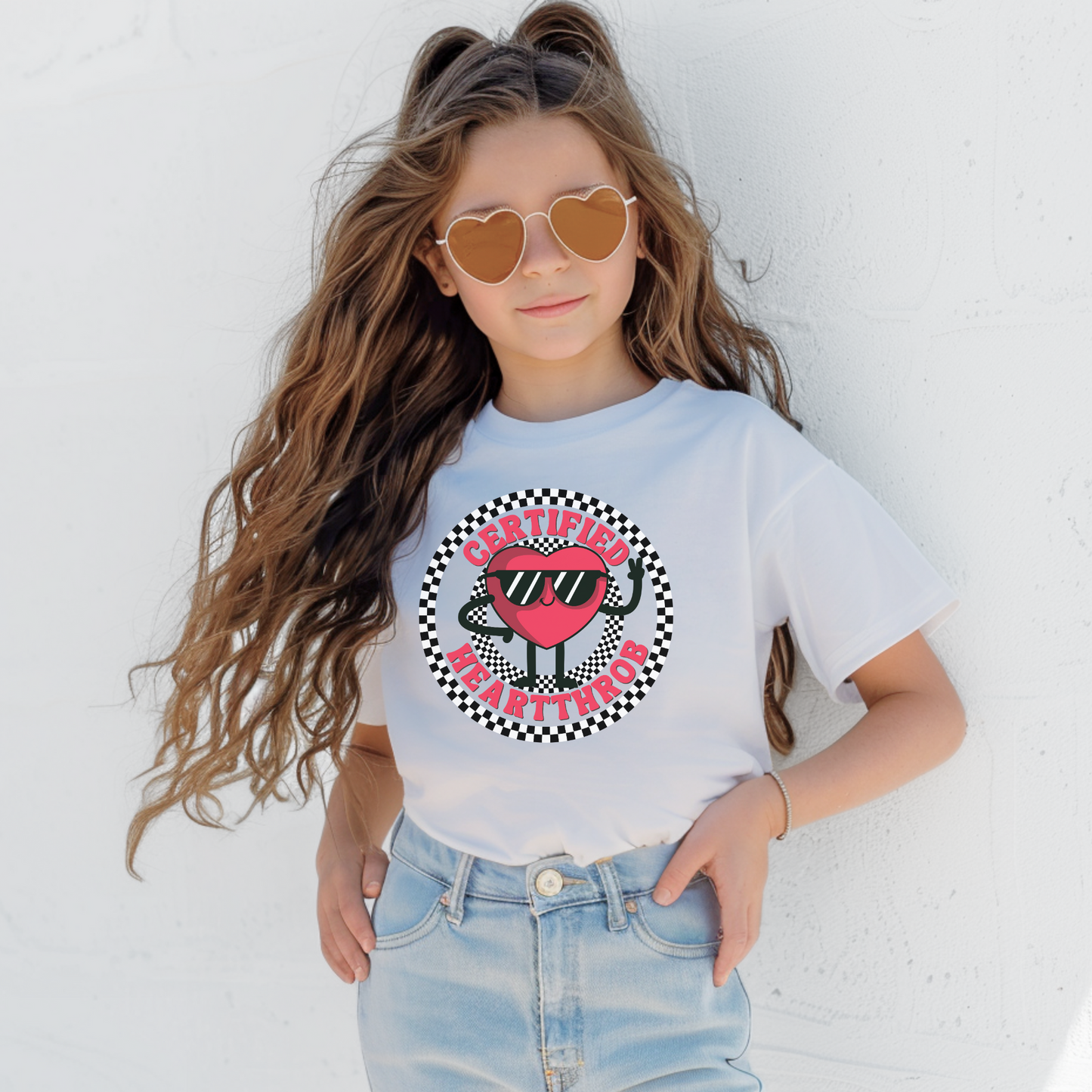 Child wearing a white Certified Heartthrob T shirt with a colorful cartoon heart wearing sunglasses at the center. The illustration is framed by a bold circular checkerboard pattern in black and white, with playful lettering around the heart. This fun youth shirt makes a cheerful addition to a Valentine's Day wardrobe.